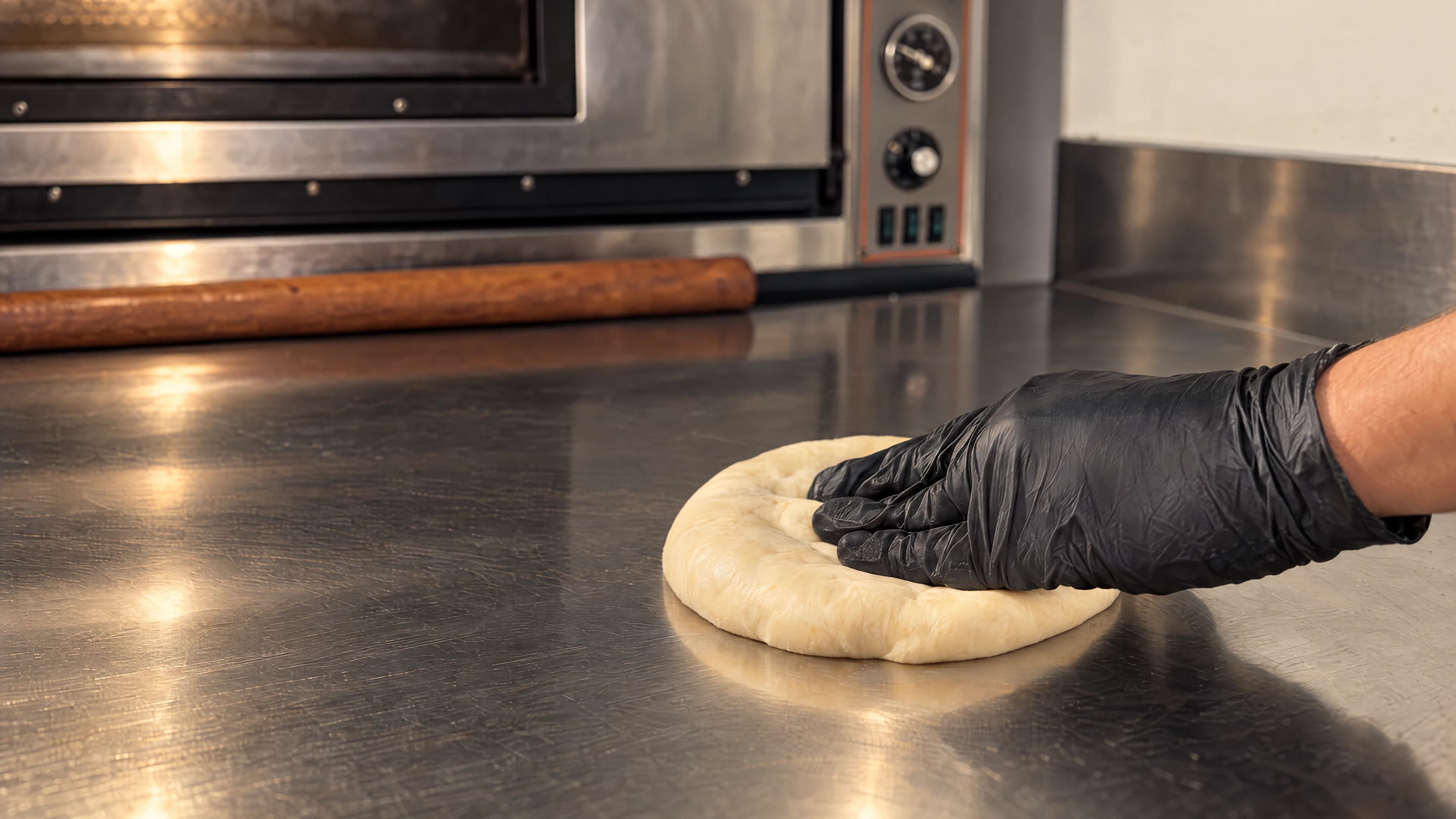 Main gantée pressant une pâte à naan sur un plan de travail en inox, devant le four.